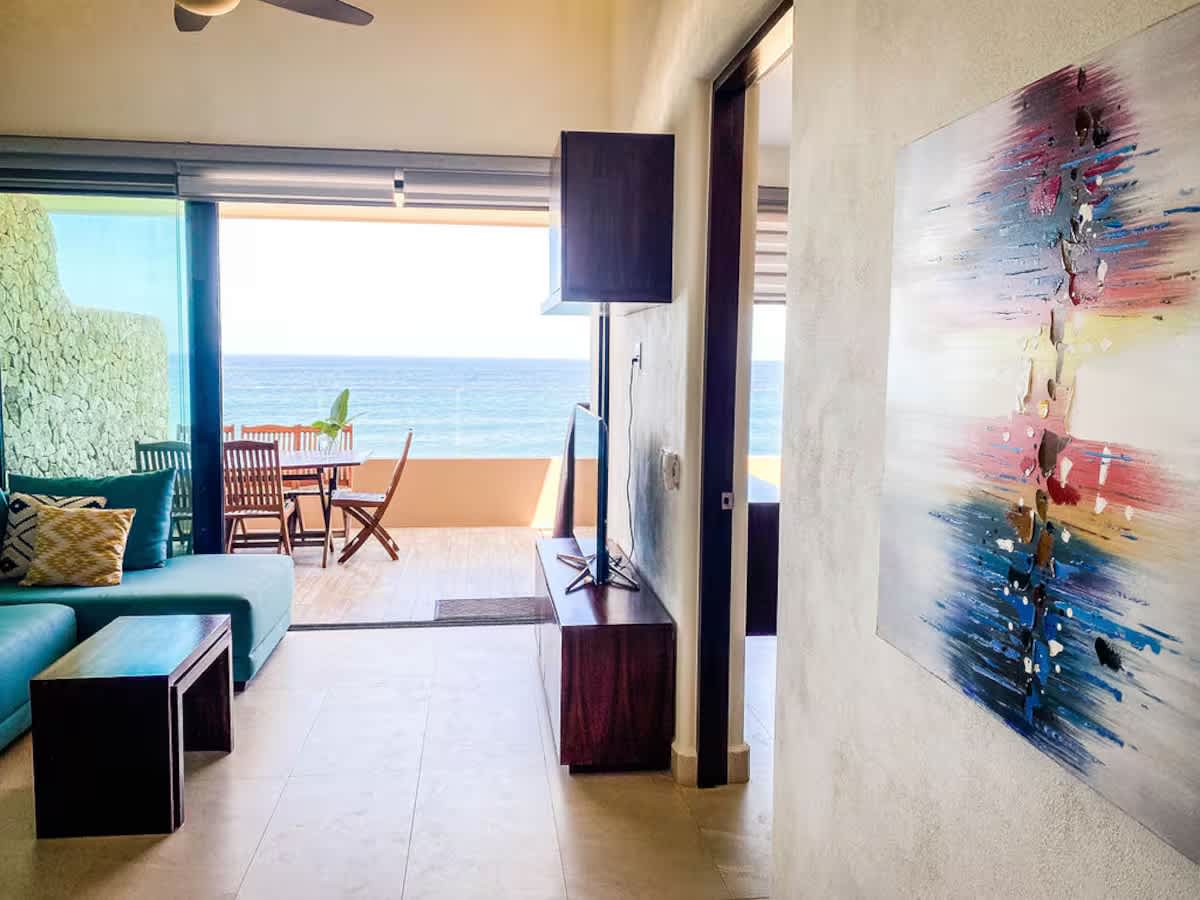 A contemporary living room with a wall-mounted TV above a dark wood console, a large abstract painting on the wall, and a view towards the ocean through a sliding glass door.