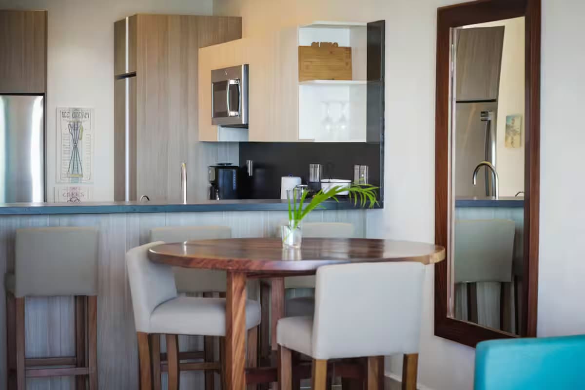 Contemporary kitchen and dining space with light wood cabinets, stainless steel microwave, a dark backsplash, and a breakfast bar with three upholstered stools, next to a round wooden dining table.
