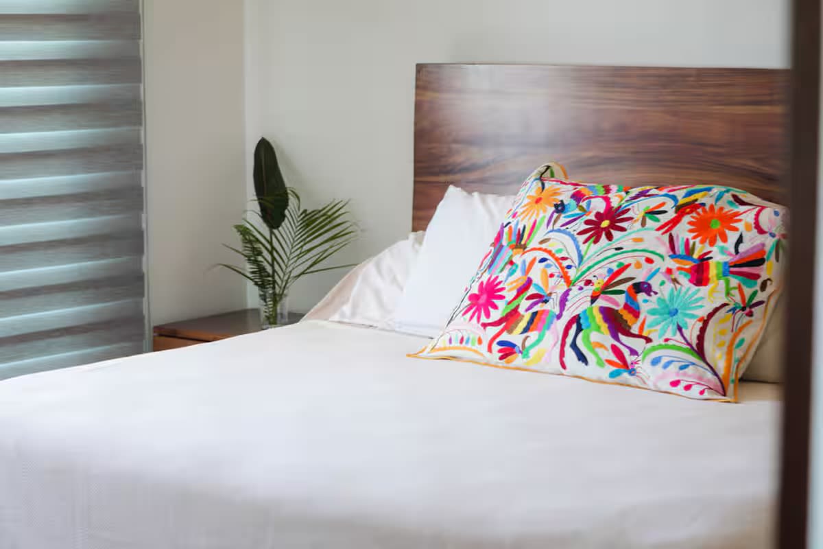 Cozy bedroom featuring a bed with crisp white linens and a vibrant, embroidered decorative pillow, complemented by a dark wooden headboard and a window with blinds.