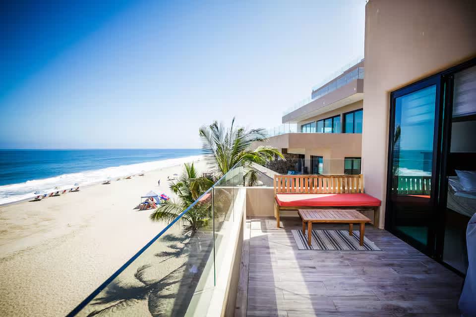 Expansive private balcony with a glass railing, offering panoramic views of a sandy beach and the vast blue ocean, furnished with a wooden bench and coffee table.