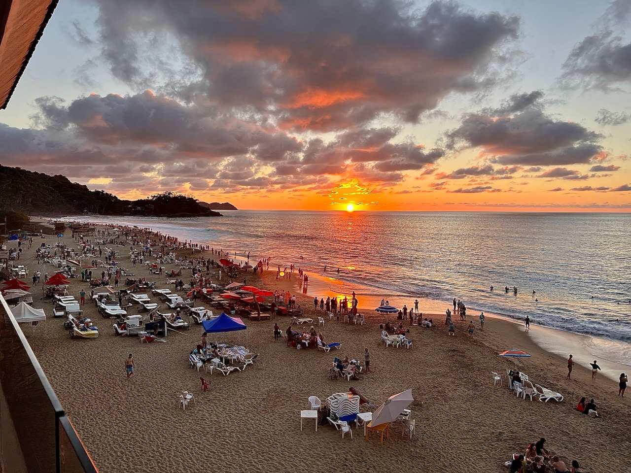 A breathtaking sunset view from an elevated perspective, showcasing a wide sandy beach bustling with people, umbrellas, and lounge chairs. The sun, a vibrant orange orb, dips into the ocean, casting a fiery glow across the water and dramatic, cloud-filled sky.
