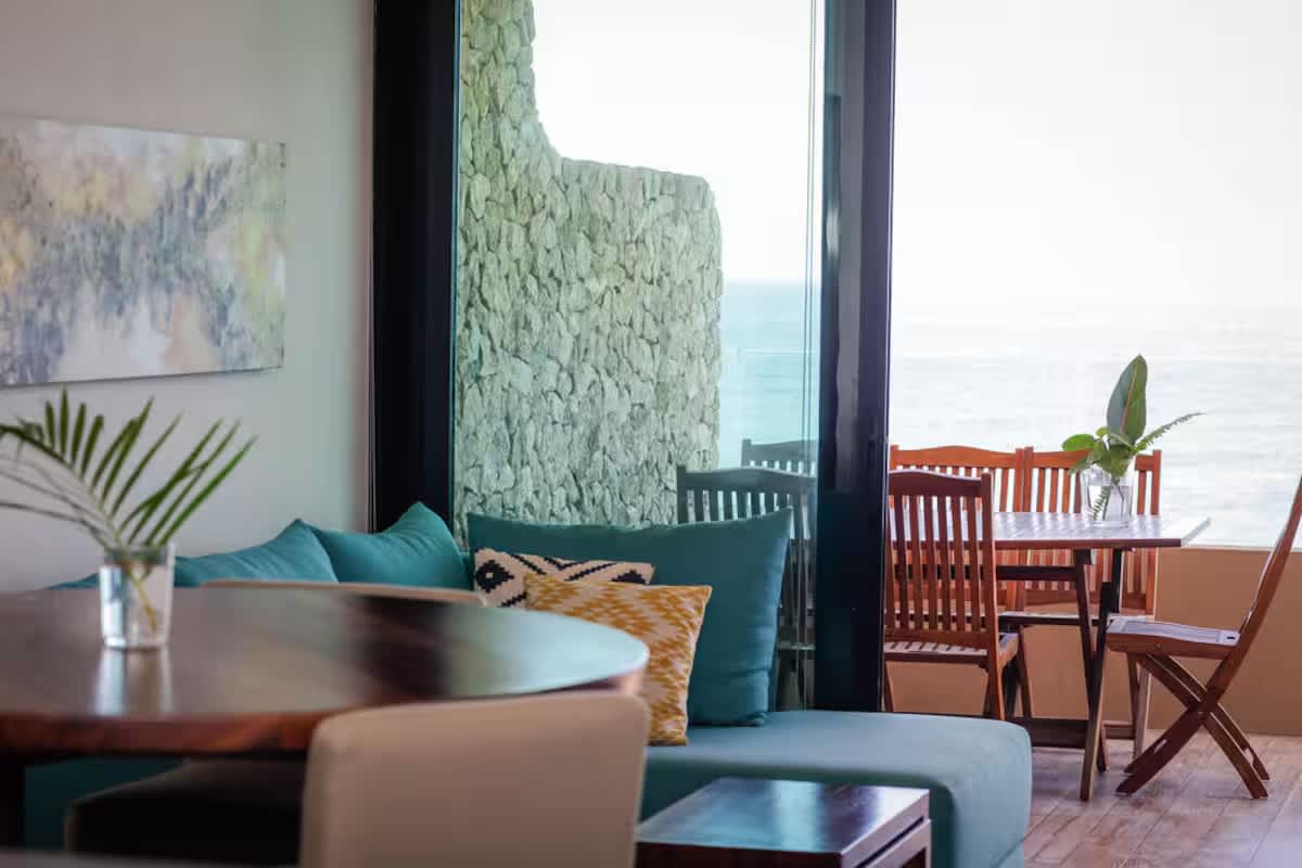 Bright living area with a teal sectional sofa and decorative pillows, a round wooden dining table, and large glass doors opening to a balcony with a stone wall and ocean view.