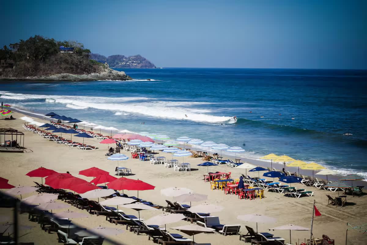 A wide shot of a vibrant sandy beach lined with numerous colorful umbrellas and lounge chairs, with gentle waves rolling into the shore and distant hills under a clear blue sky.