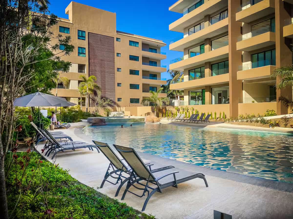 Expansive resort swimming pool with a large deck area and numerous lounge chairs, flanked by modern multi-story buildings and vibrant green landscaping under a clear blue sky.