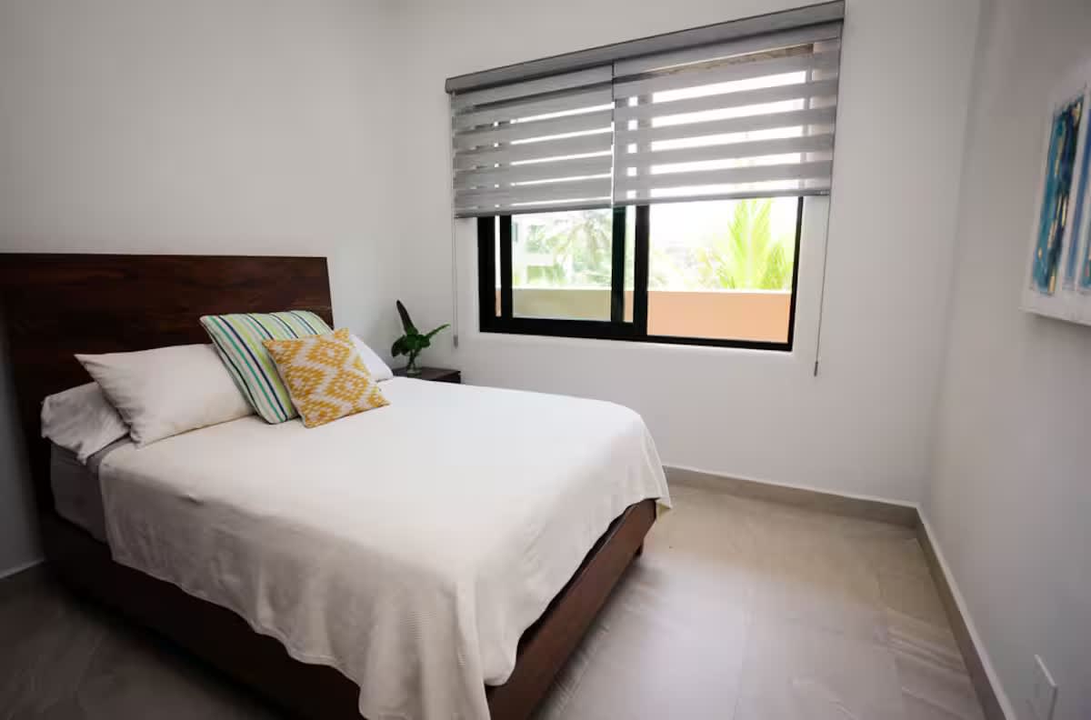 A bright bedroom with a dark wood bed frame and white bedding, featuring a large window with horizontal blinds that offers a glimpse of greenery outside.