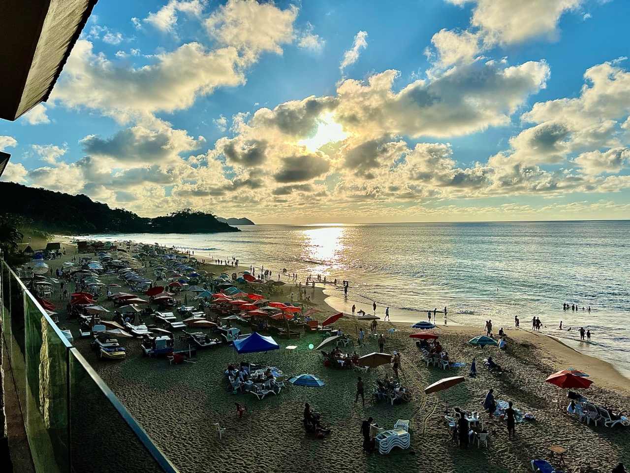 A vibrant daytime view from an elevated position, overlooking a sandy beach filled with people, colorful umbrellas, and lounge chairs. The bright sun shines through scattered clouds, creating a sparkling reflection on the calm blue ocean.