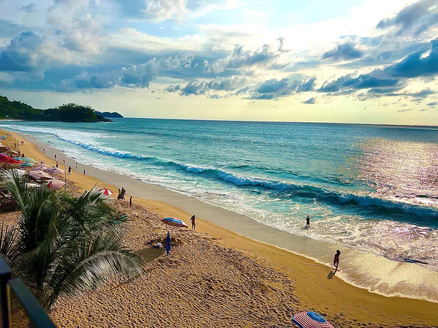 Wide sandy beach with gentle ocean waves under a partly cloudy sky, dotted with beach umbrellas and people enjoying the water and sun.