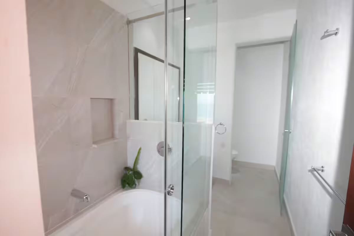 Sleek bathroom with a white bathtub enclosed by clear glass panels, featuring modern chrome fixtures and light-colored tiled walls, with a view into the toilet area.
