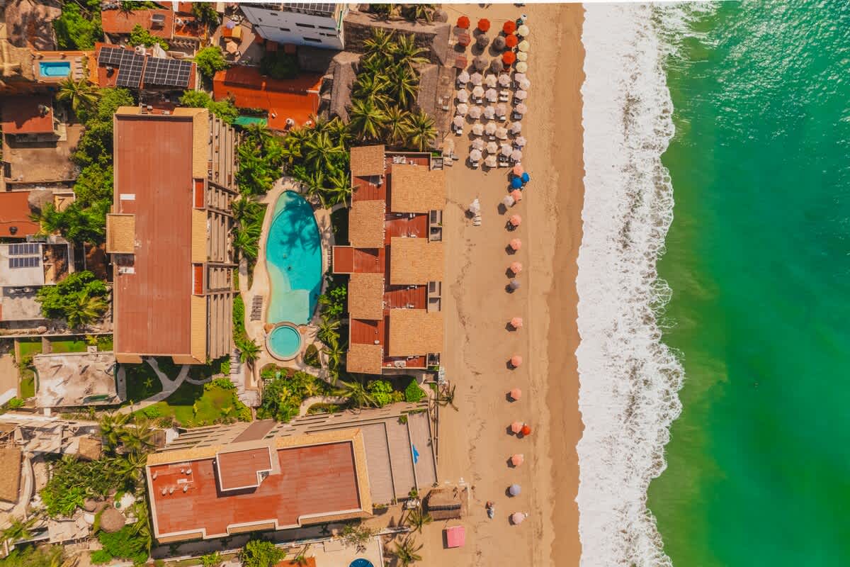 An aerial view showcases the beachfront property with multiple buildings, a large swimming pool, lush green landscaping, and a wide sandy beach with umbrellas bordering the turquoise ocean.