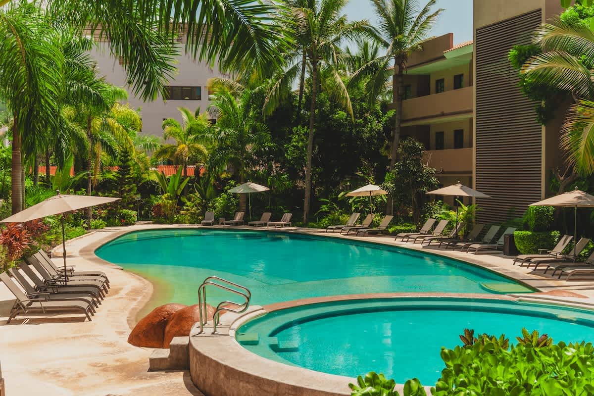 A large, freeform swimming pool with a connected circular hot tub, surrounded by numerous lounge chairs with umbrellas on a concrete deck. Lush tropical landscaping, including tall palm trees, frames the pool area, with multi-story buildings in the background.