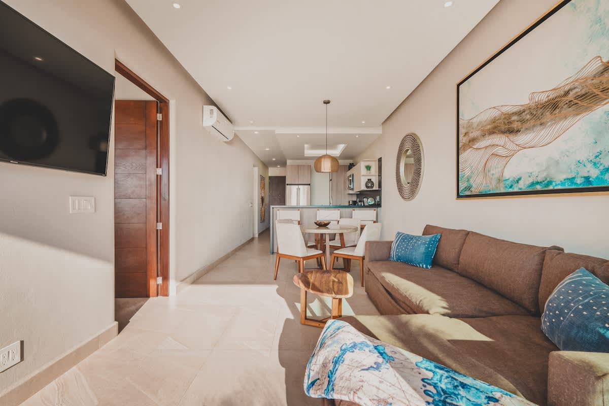 Open-concept living room with a large brown sectional sofa, a round dining table with four chairs, and a flat-screen TV, leading to the kitchen and offering a bright, airy feel.