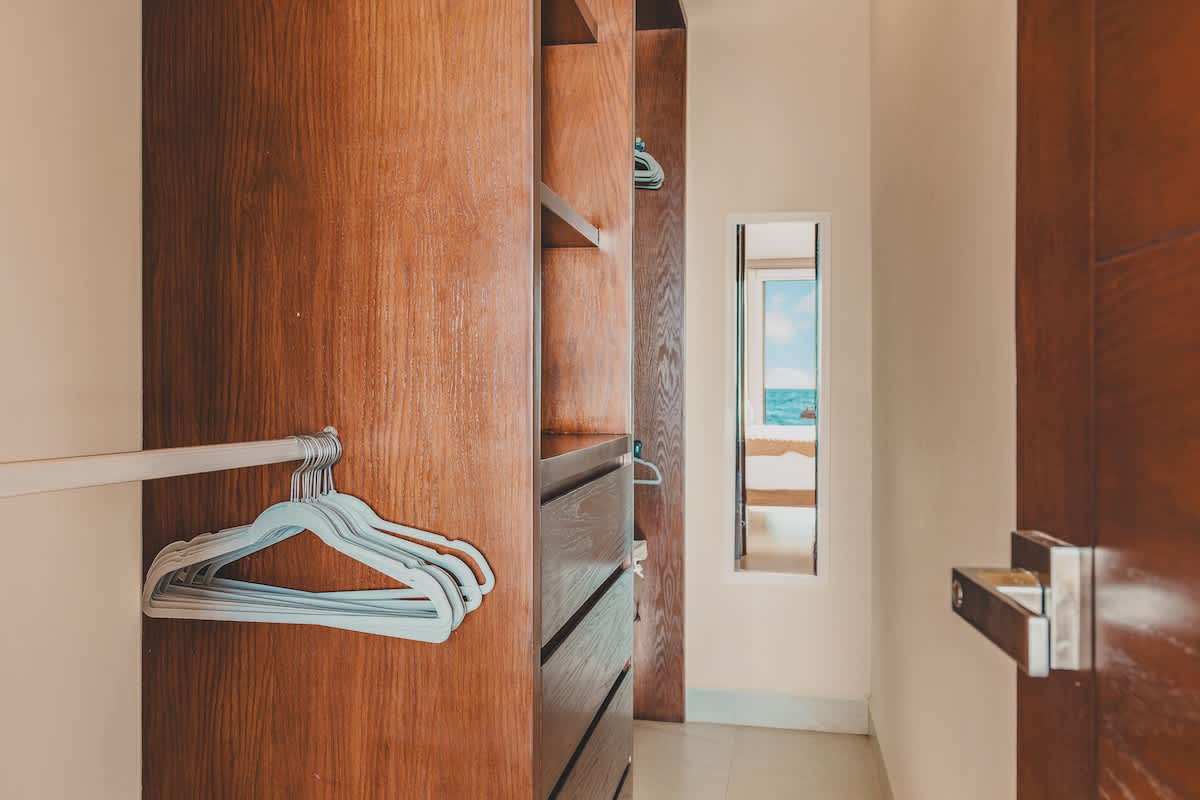 A spacious walk-in closet features dark wood shelving units, drawers, and hanging rods with several white hangers, reflecting a glimpse of a bedroom and ocean view in the mirror.