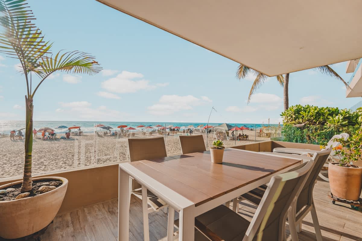 A spacious beachfront balcony features an outdoor dining table with six chairs, set on a wooden-look tiled floor. A small potted palm tree adds greenery, and the view overlooks a sandy beach with numerous umbrellas and the ocean.