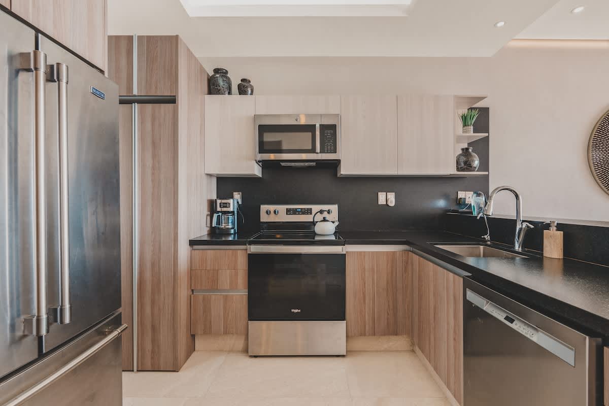 Bright kitchen with light wood cabinets, dark countertops, and stainless steel appliances including a side-by-side refrigerator, microwave, and electric stove, complemented by decorative vases on open shelving.