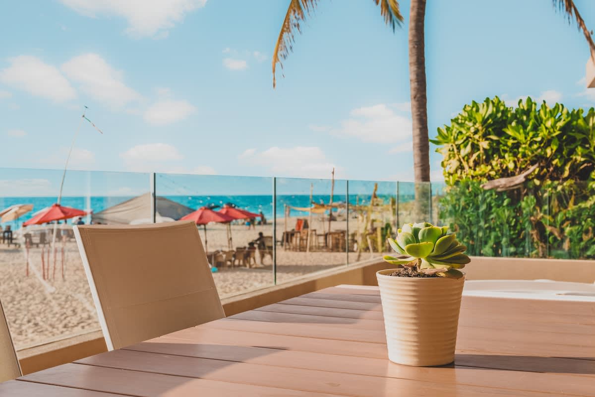 View from an outdoor dining table on a balcony, featuring a small potted succulent, with the sandy beach, ocean, and distant beach umbrellas visible through a clear glass railing.