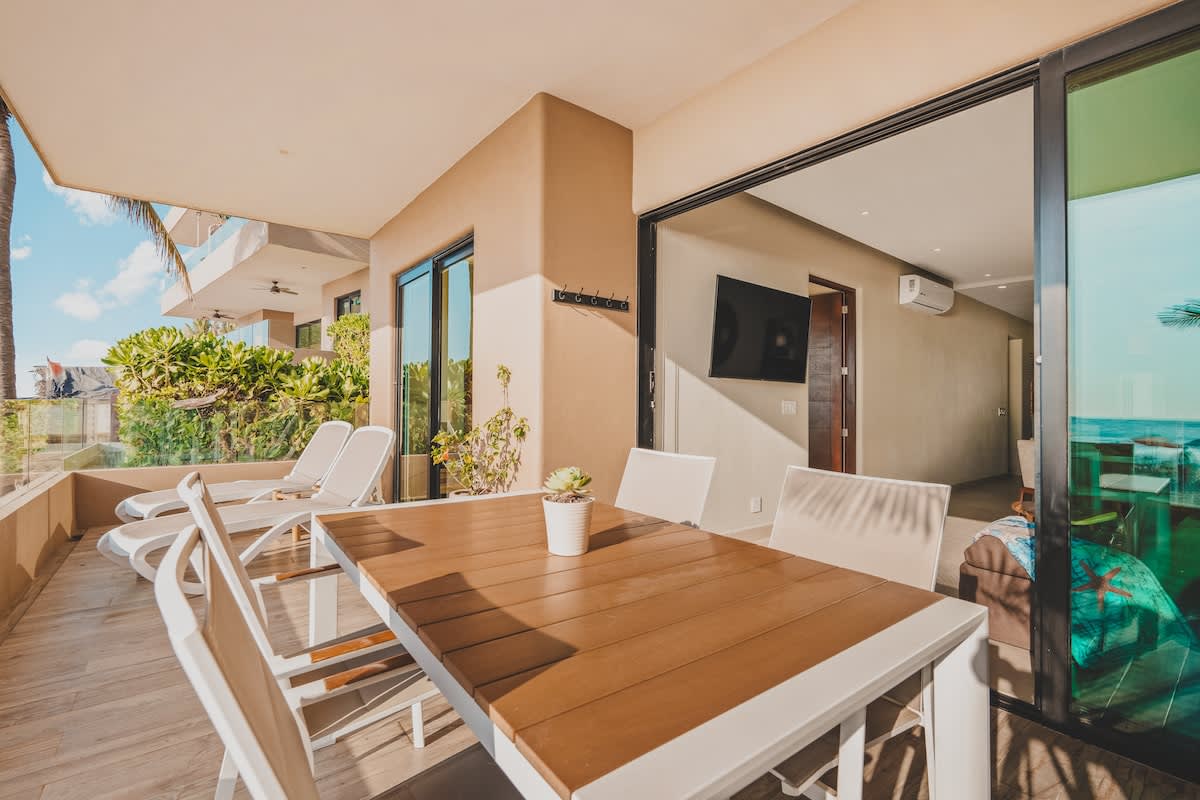 Spacious beachfront balcony with a wooden-topped outdoor dining table and chairs, two lounge chairs, and a stone accent wall, offering views of the ocean and adjacent properties.