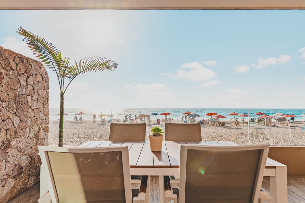 Close-up view from a beachfront balcony, featuring an outdoor dining table with a small potted plant, overlooking a sandy beach bustling with umbrellas and people, with the ocean stretching to the horizon.