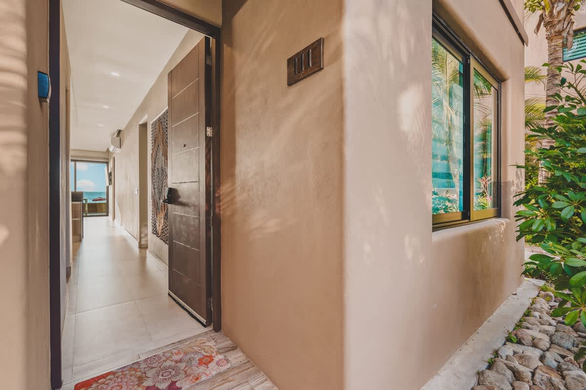 The property entrance features a dark wooden door, light beige stucco walls, and a hallway with tiled flooring leading towards the interior, with a glimpse of the ocean in the distance.