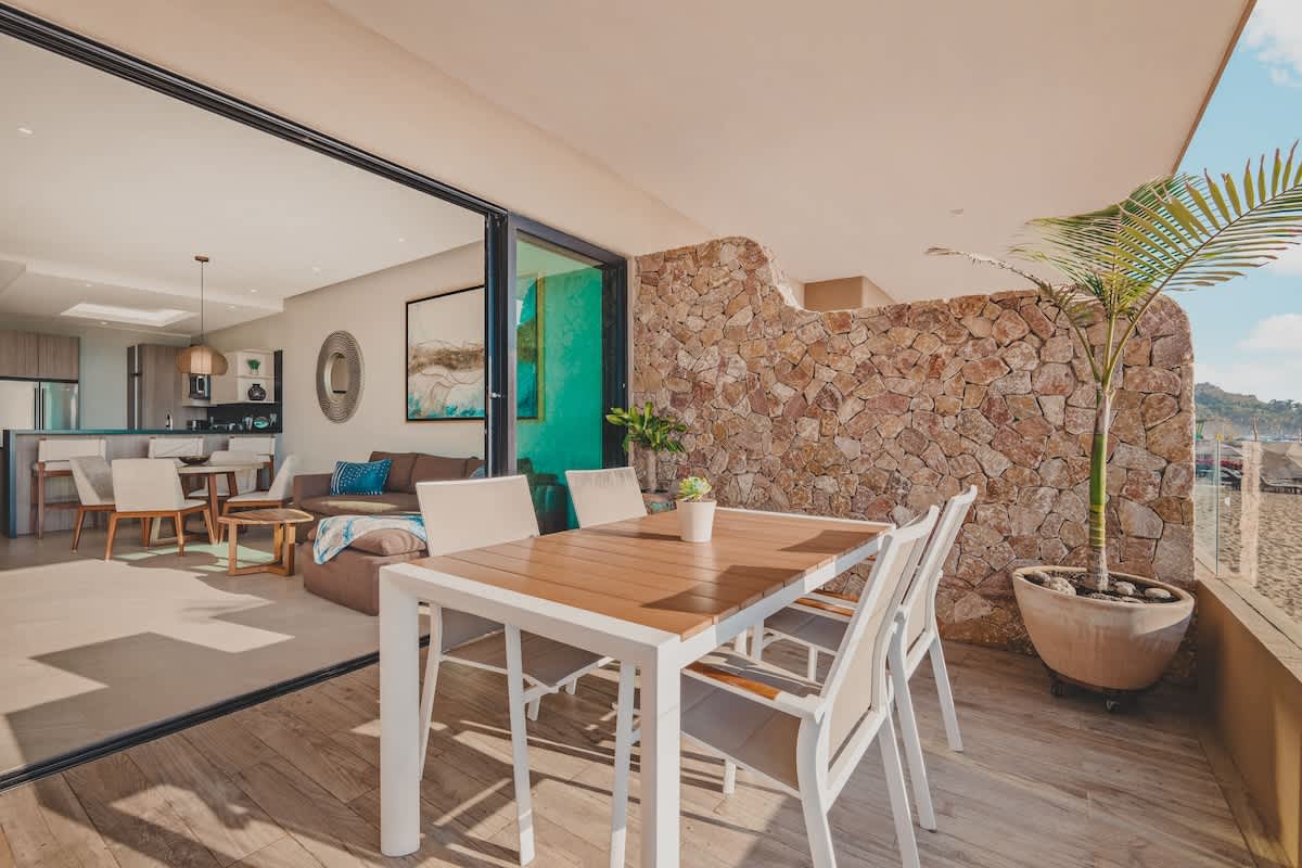 Wide shot of a beachfront balcony with an outdoor dining set and a stone accent wall, looking into the open-concept living area with a sofa and kitchen beyond through large sliding glass doors.