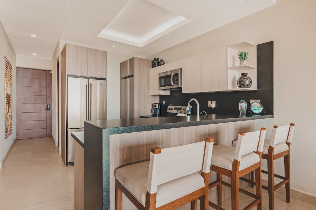 Kitchen island with dark countertops and three bar stools, providing a casual dining space, with a view towards the main entrance door and the stainless steel refrigerator.