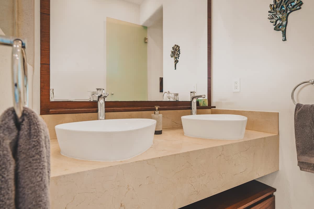 A close-up of a modern double vanity featuring two white oval vessel sinks on a light-colored stone countertop. A large mirror with a dark wood frame is mounted above.