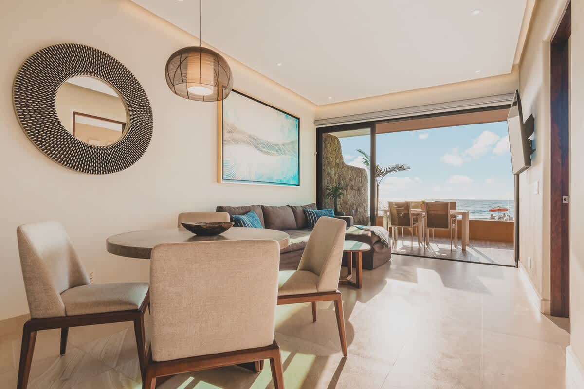 An open-concept living and dining area features a round wooden dining table with four upholstered chairs, a large circular mirror, and a brown sectional sofa in the background with ocean views.