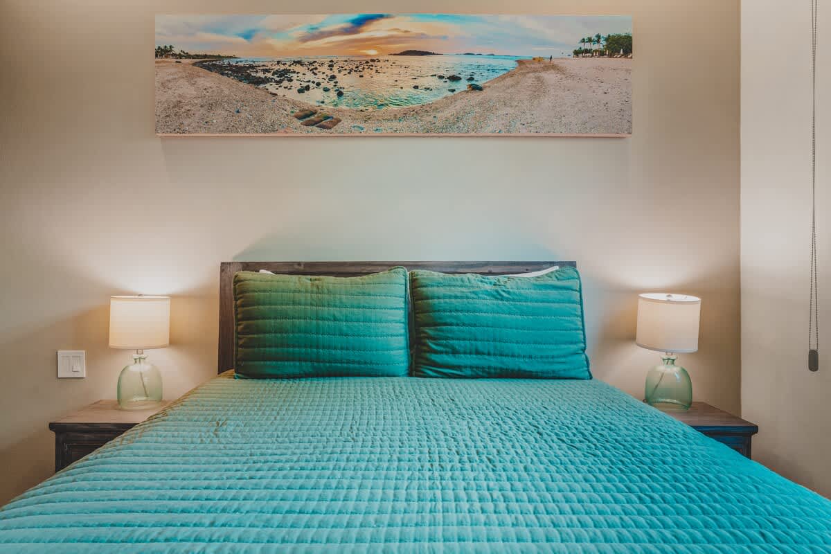 A closer view of Bedroom-2's bed, showcasing the teal quilted bedding and pillows, flanked by two rustic wooden nightstands with glass lamps. A panoramic beach scene artwork is mounted above the bed.