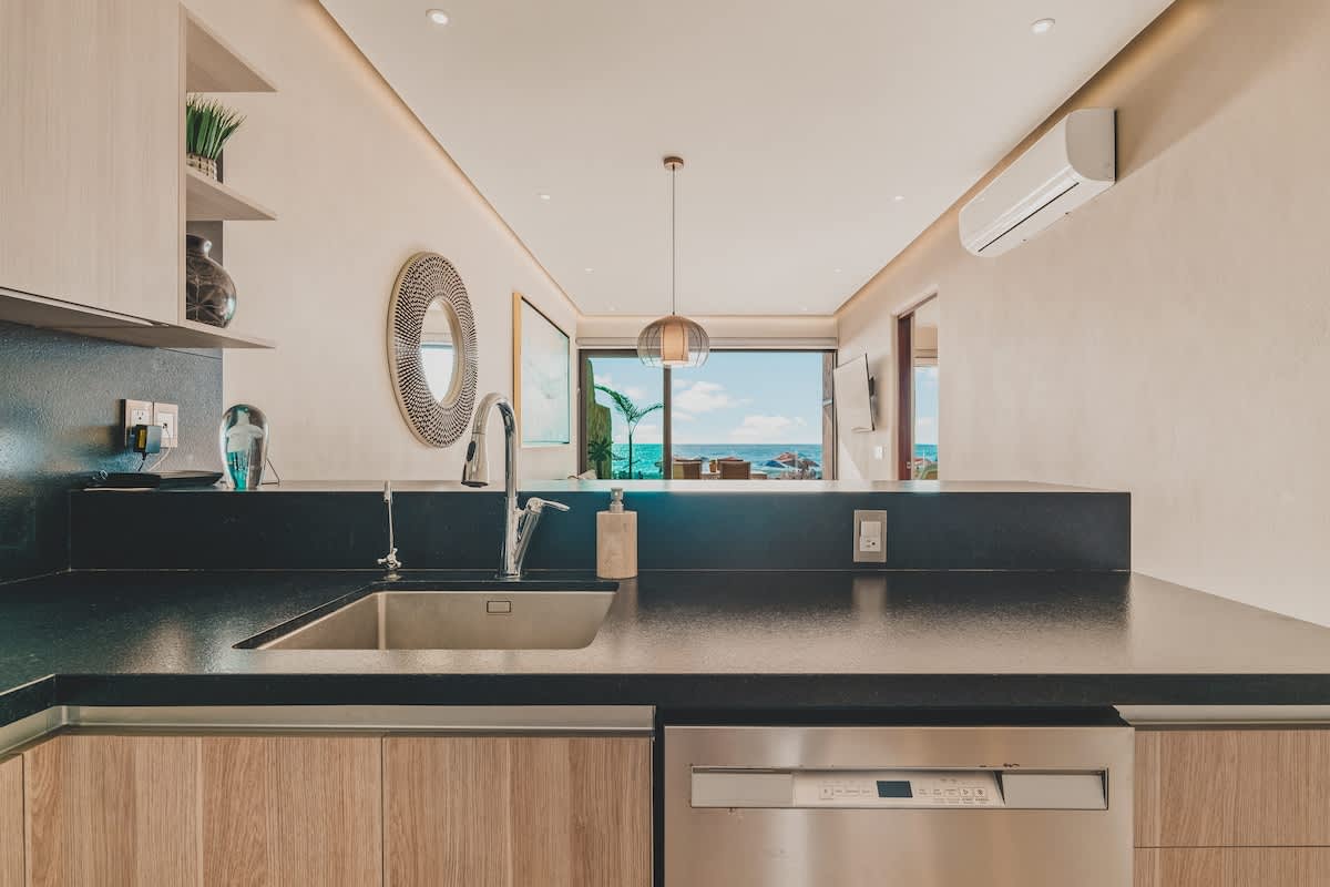 Close-up of the modern kitchen counter featuring a stainless steel sink with a gooseneck faucet, dark countertops, light wood base cabinets, and a glimpse of the ocean view through the living area.