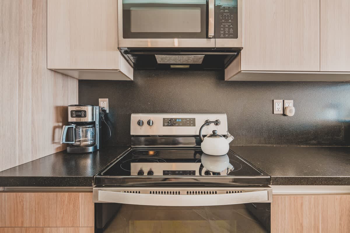 Close-up of the kitchen's stainless steel electric stove with a white kettle, a coffee maker, and an over-the-range microwave, set against a dark backsplash and light wood cabinets.