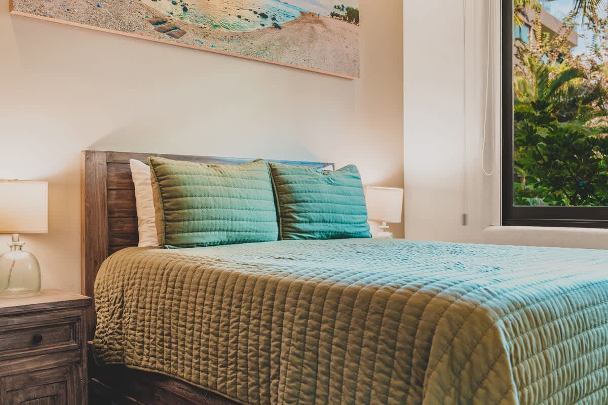 A side view of Bedroom-2's bed, featuring a teal quilted comforter and pillows, with a dark wooden headboard. A window to the right provides a glimpse of vibrant green tropical plants, enhancing the room's natural ambiance.