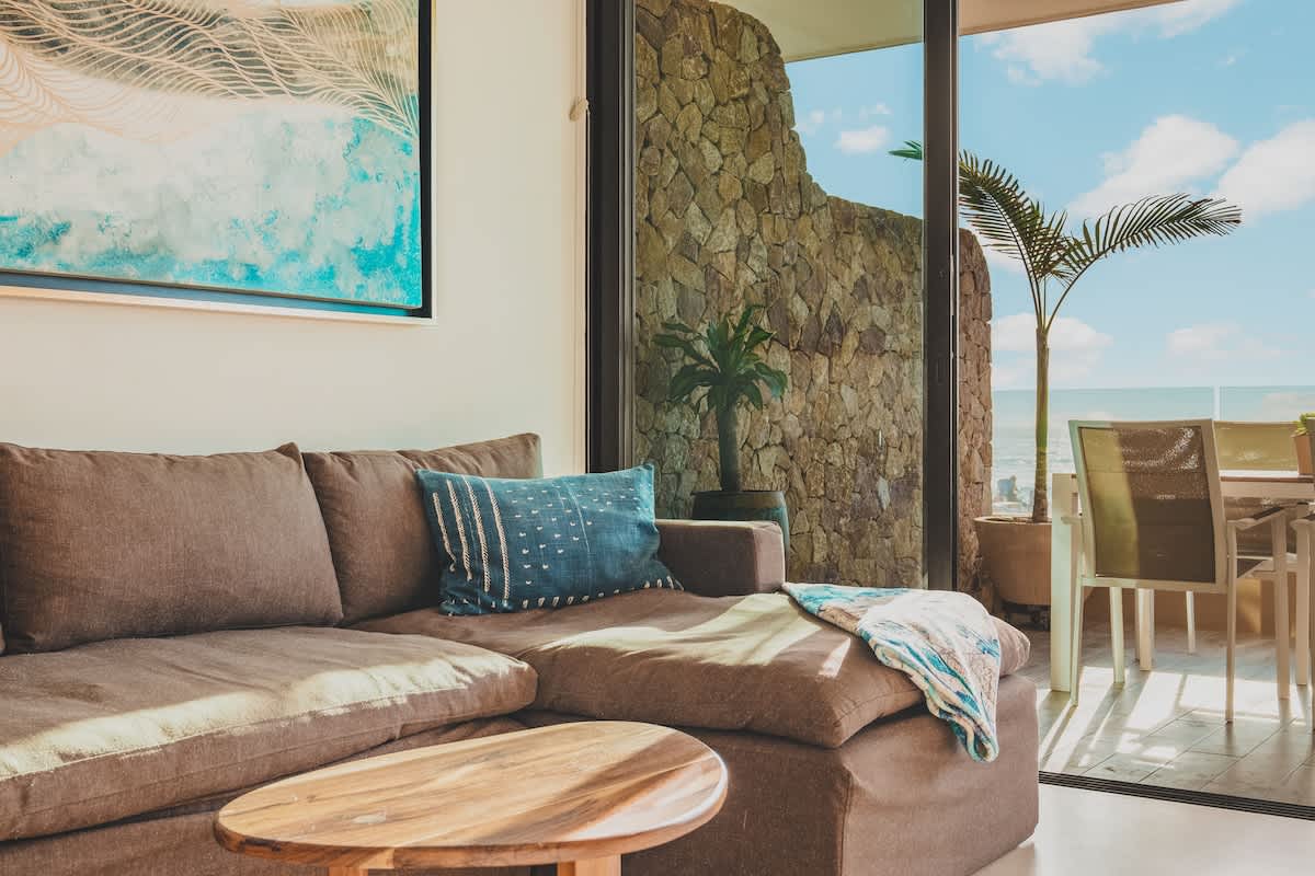 A cozy living room features a brown sectional sofa with a blue patterned throw pillow, a wooden oval coffee table, and large glass doors opening to an outdoor patio with ocean views.