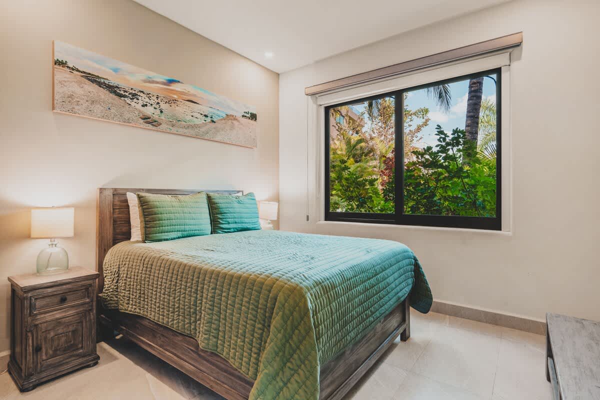 A cozy bedroom with light walls, a wooden bed frame, and a teal quilted bedspread. A large window offers a lush green view, and a long landscape painting hangs above the bed.
