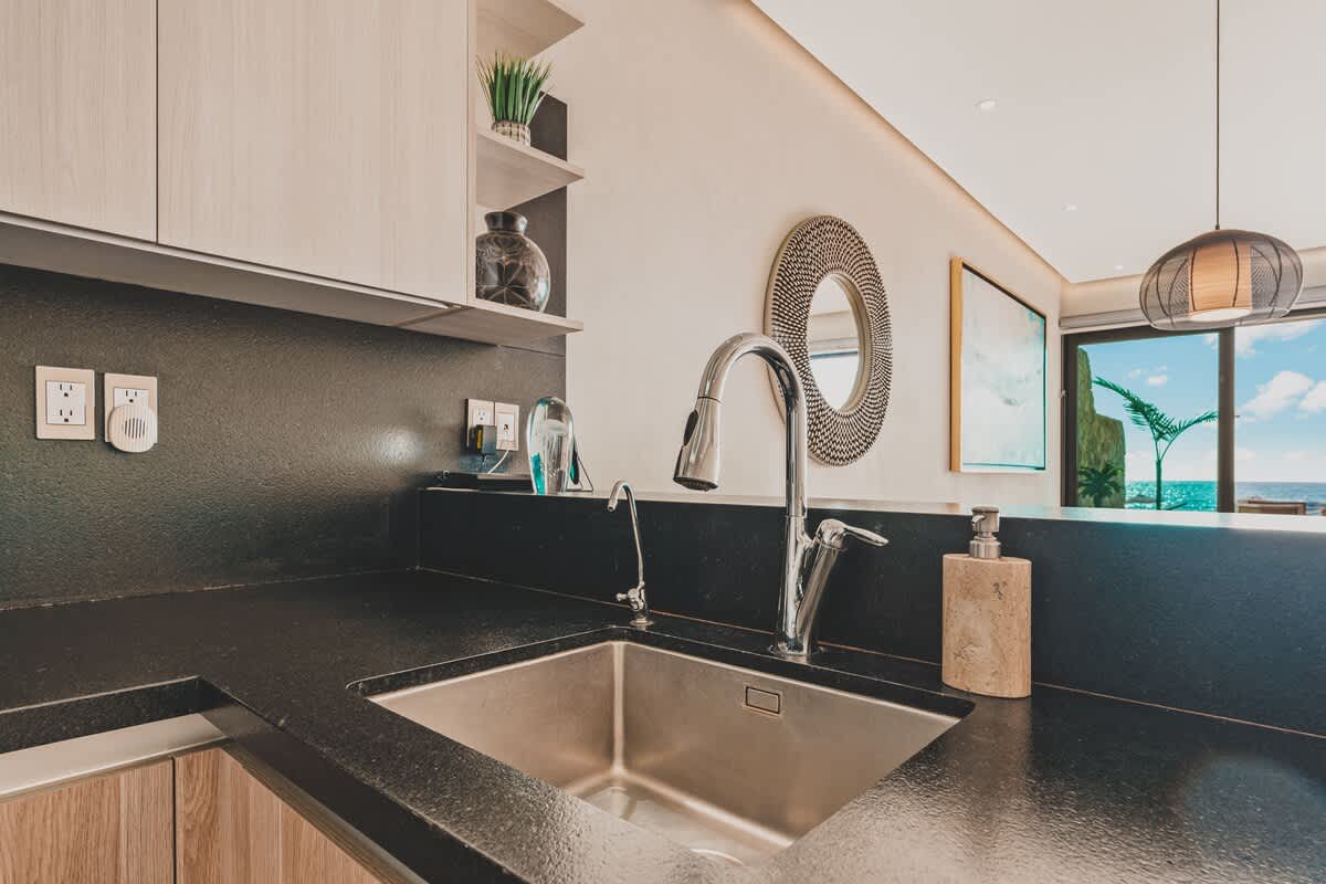 Detailed view of the kitchen sink area with a modern gooseneck faucet, dark countertops, light wood cabinets, and a decorative round mirror on the wall, enhancing the contemporary design.