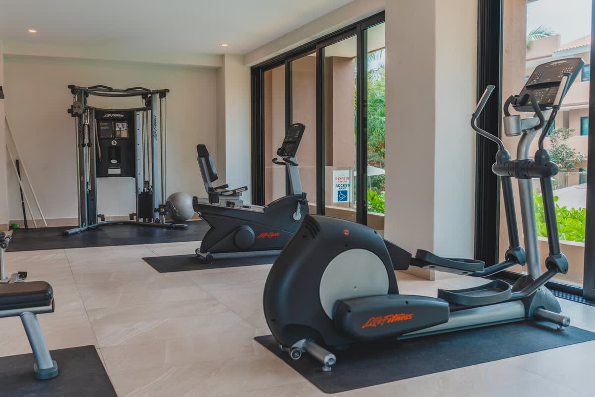 A bright gym space features various cardio machines including an elliptical and stationary bikes, with large windows offering views of the outdoors and tiled flooring.