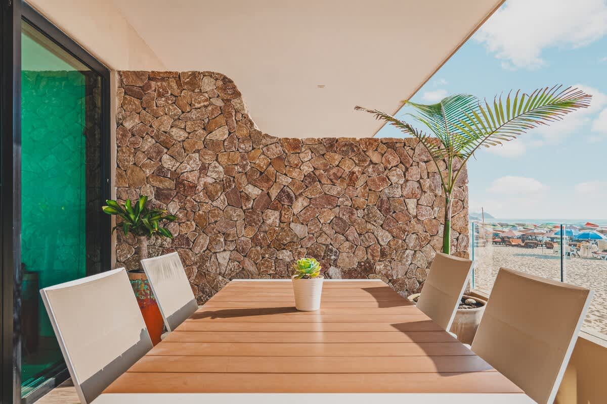 Outdoor dining area on a balcony with a wooden-topped table and chairs, set against a textured stone wall and a potted palm tree, with a glimpse of the beach and ocean in the background.