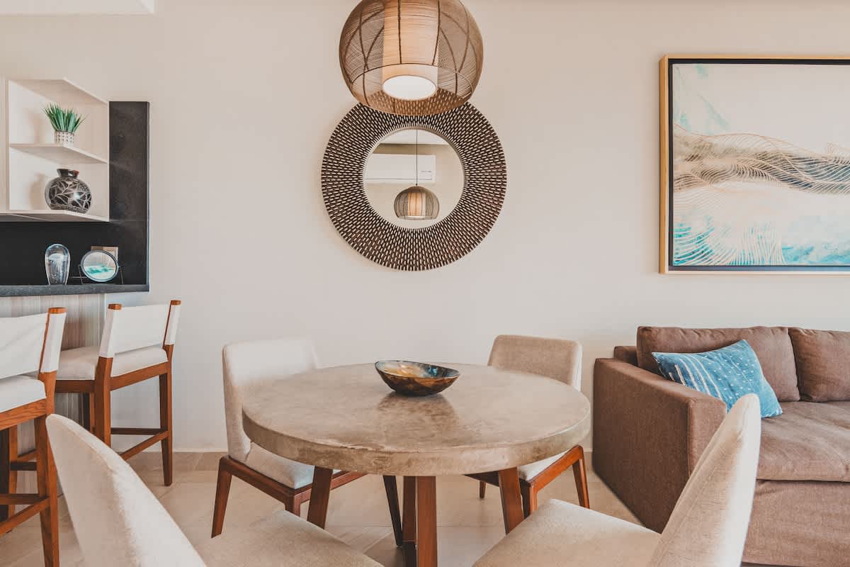 Modern dining area with a round concrete-like table and four light-colored chairs, illuminated by two woven pendant lights, adjacent to a living area with a brown sofa and a decorative mirror.