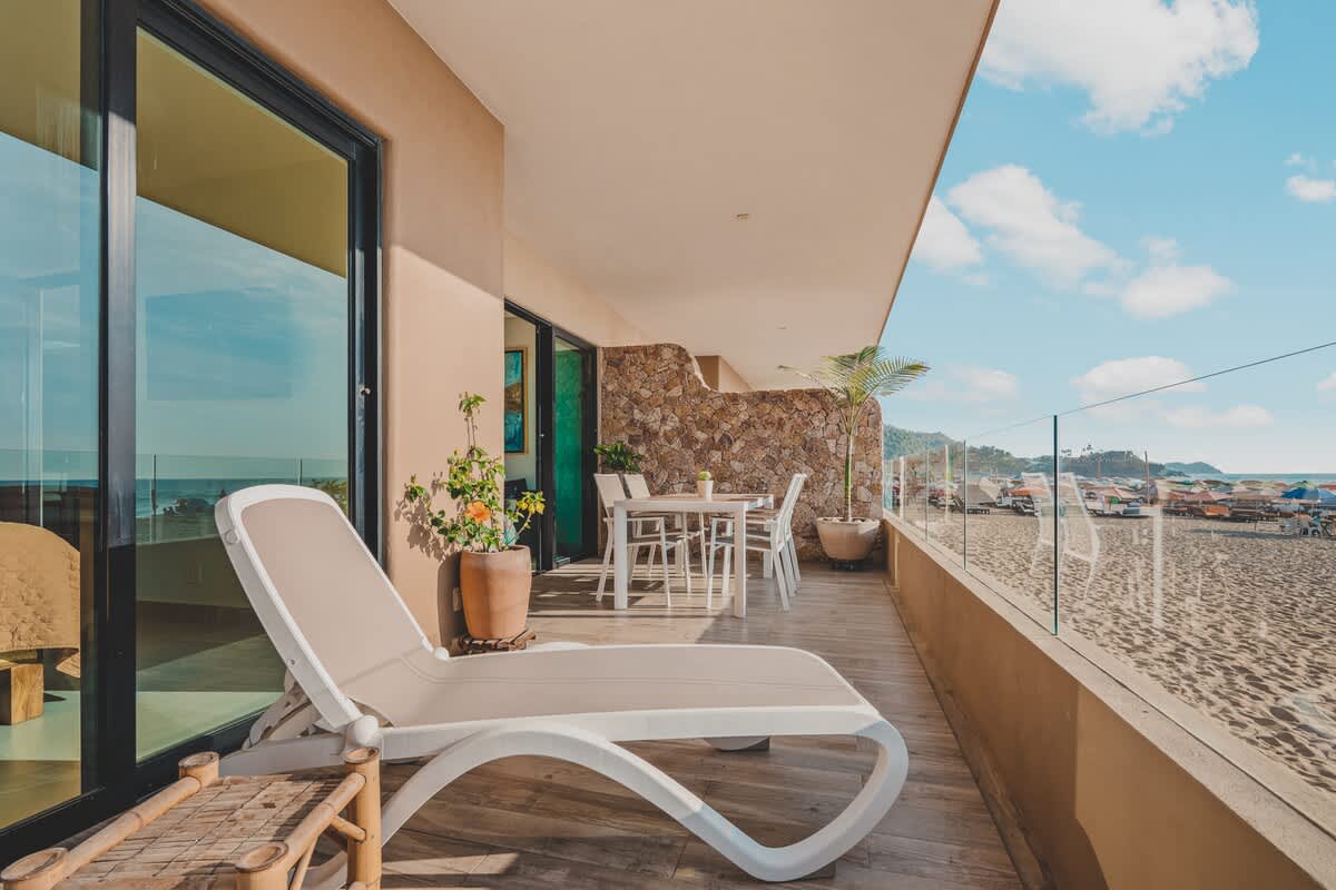 Expansive beachfront balcony with a mix of lounge chairs and an outdoor dining set, featuring a glass railing that provides panoramic views of the sandy beach and the ocean.