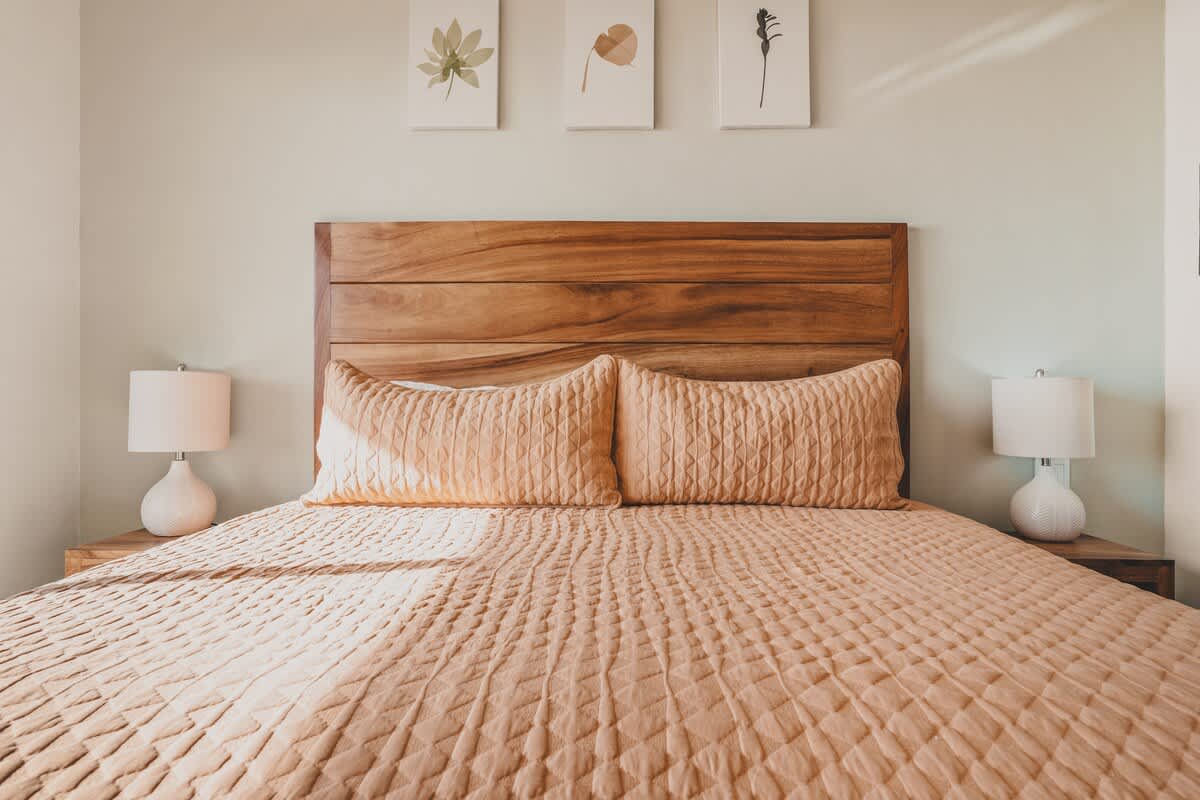 A bright bedroom centered around a large wooden headboard and a bed with a light peach quilted bedspread. Three minimalist botanical prints adorn the wall above the bed.