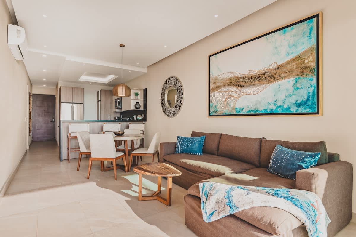 Bright and spacious living room with a brown sectional sofa, a round dining table, and a view towards the modern kitchen, featuring large abstract artwork and a decorative round mirror.
