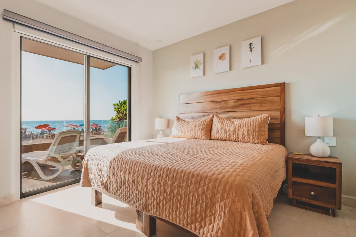A spacious bedroom with a king-sized bed and a wooden headboard, featuring a light peach quilted bedspread. Large sliding glass doors open to a balcony with ocean views and beach access.