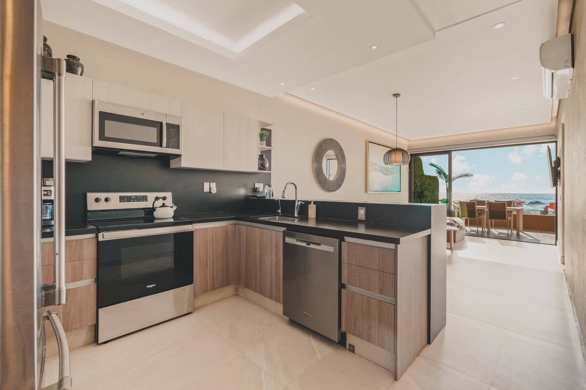 Modern kitchen with light wood cabinetry, dark countertops, stainless steel appliances including a microwave, electric stove, and dishwasher, offering an open view to the living room and ocean.