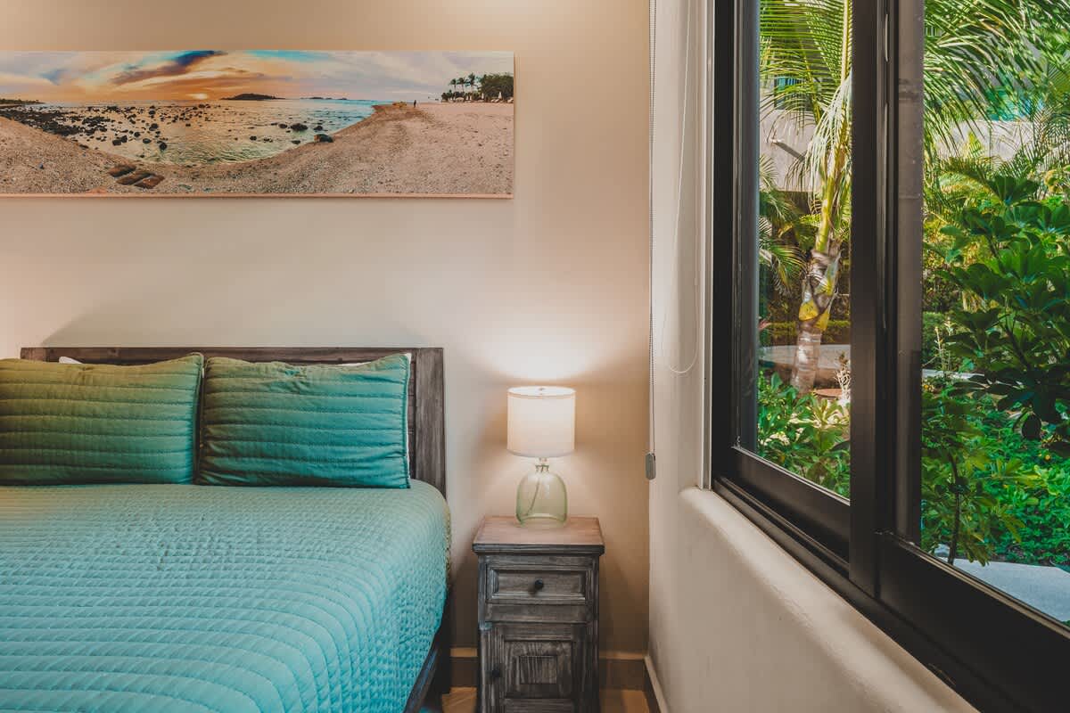 An alternative view of Bedroom-2, highlighting the bed with teal quilted bedding and a rustic wooden nightstand with a lamp. A large window offers a lush green view of tropical foliage outside.