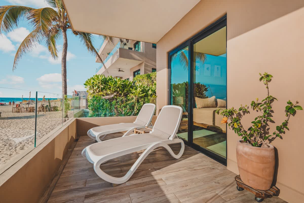 Sunny balcony featuring two modern white lounge chairs and a potted plant, with a clear glass railing offering an unobstructed view of the sandy beach and ocean.
