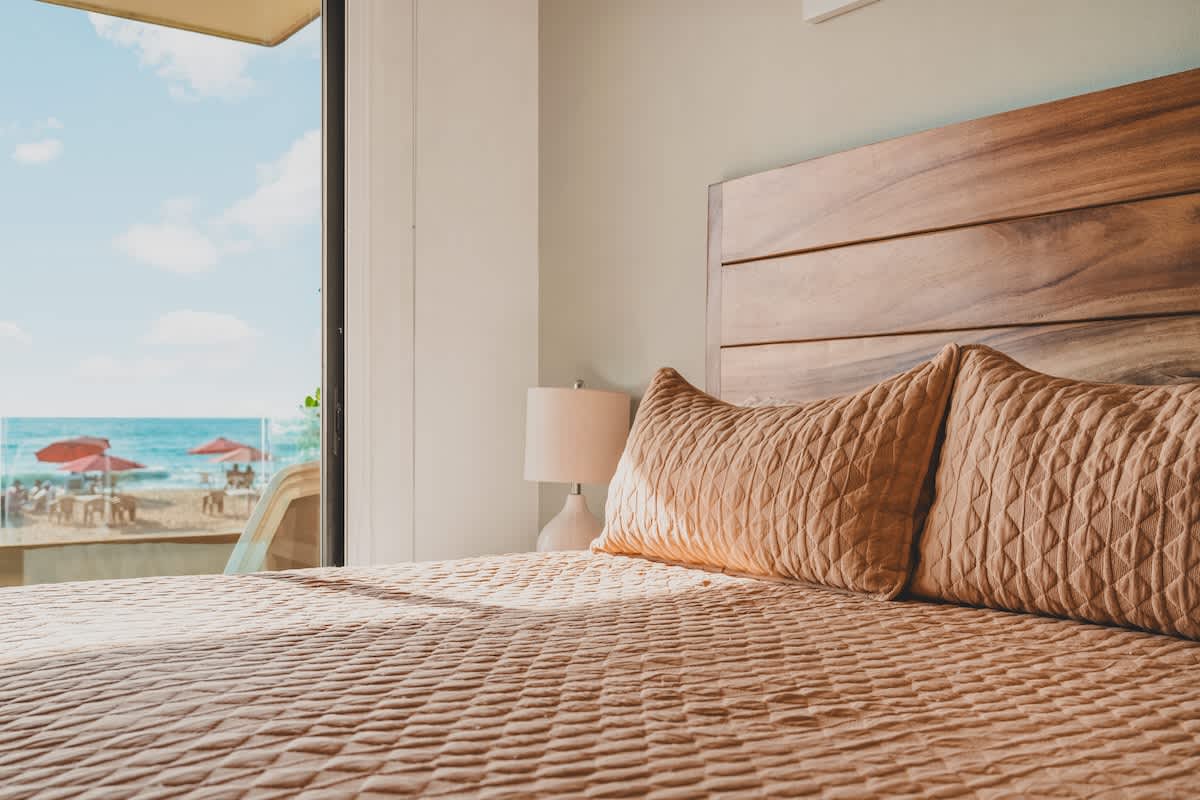 A close-up of Bedroom-1's bed, featuring peach-colored quilted bedding and pillows, with a wooden headboard. A large glass door provides a direct view of the sandy beach and ocean beyond.