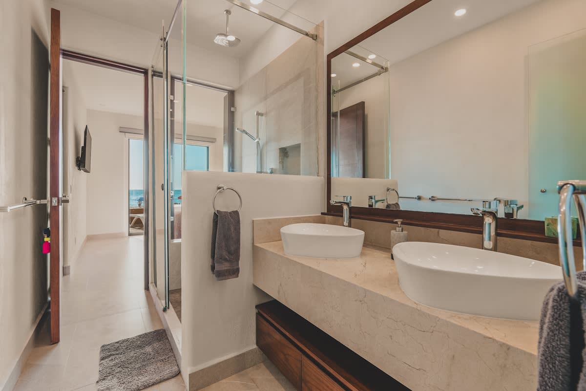 A bright bathroom featuring a double vanity with two white vessel sinks set on a light marble countertop. A large mirror with a dark wood frame hangs above, reflecting the adjacent shower and bedroom.