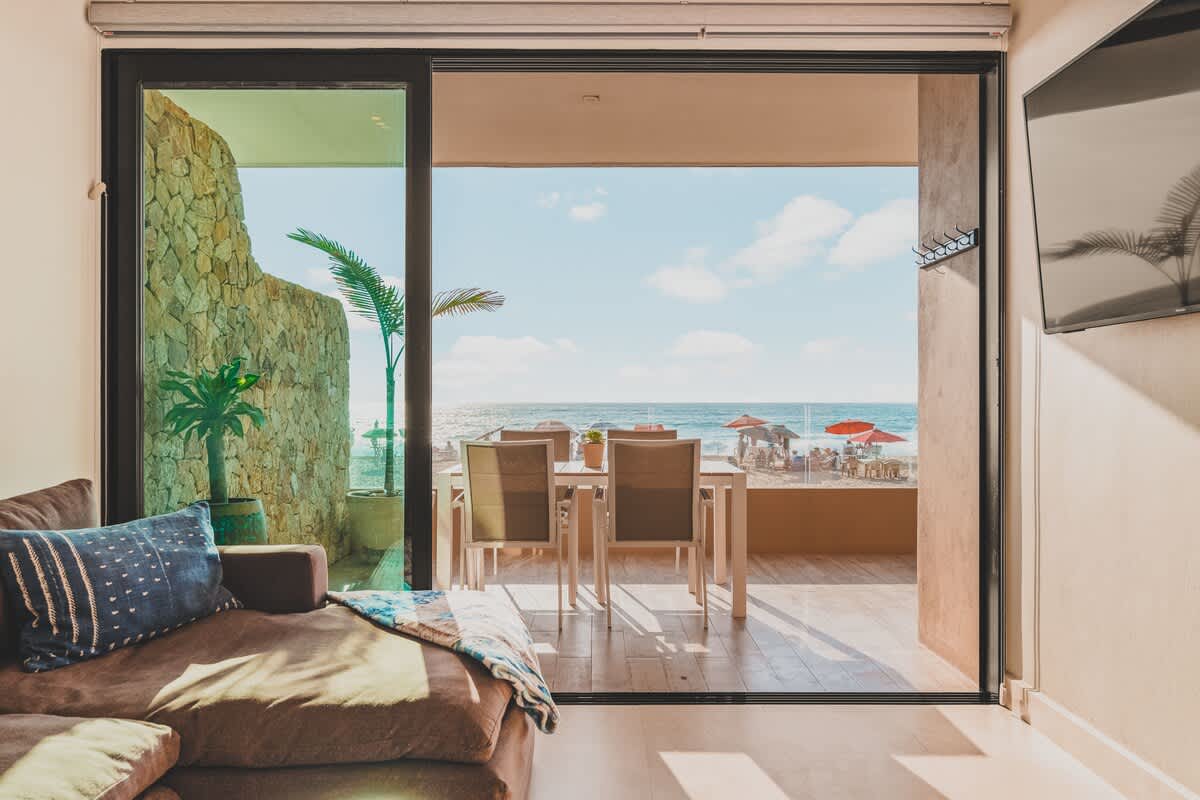 View from a cozy living room with a brown sectional sofa, looking out through large sliding glass doors onto a beachfront balcony with an outdoor dining set and a vibrant beach scene beyond.