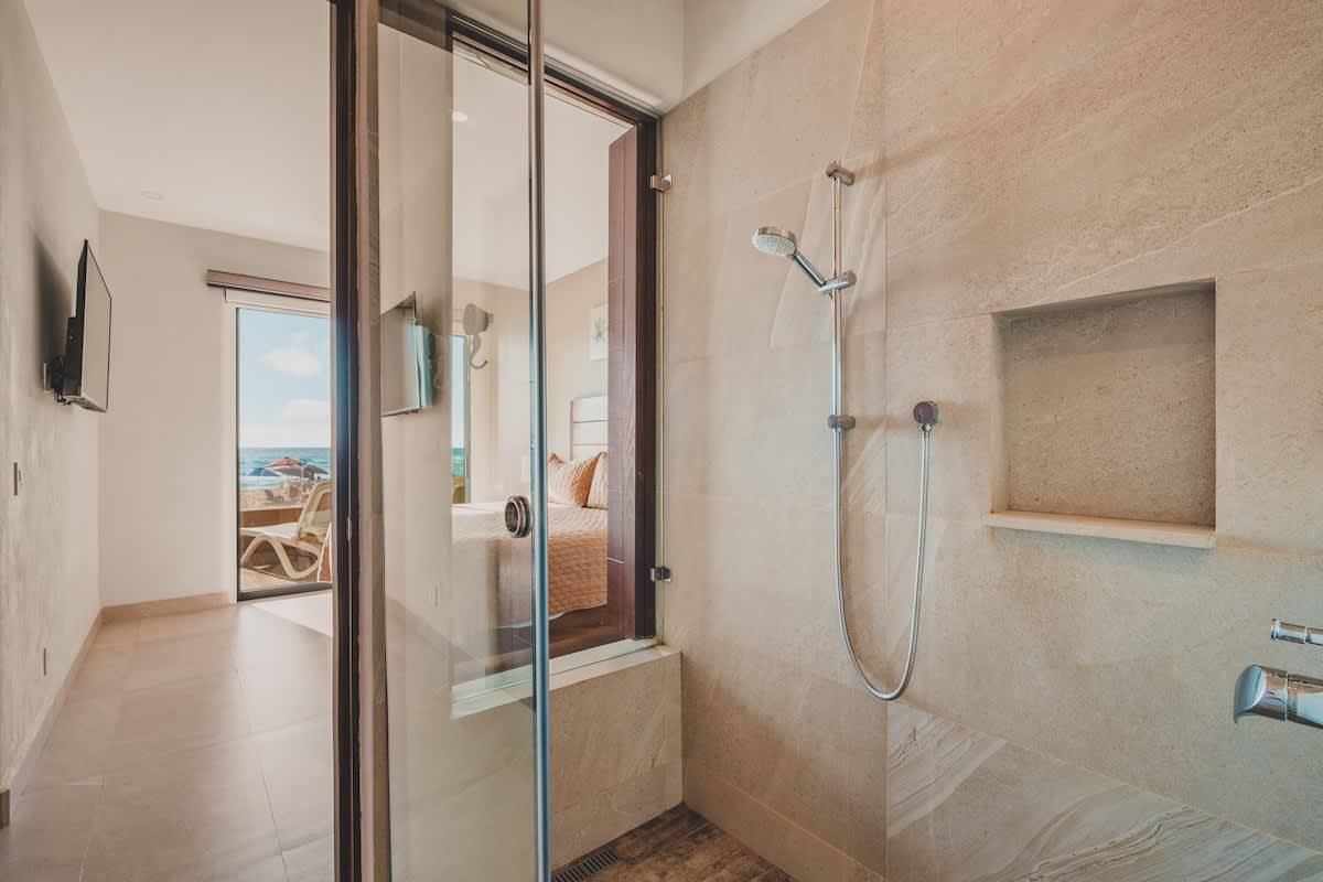 A spacious walk-in shower with light-colored tile walls and a glass enclosure, featuring a rain shower head and a built-in niche. The view extends into the adjacent bedroom.