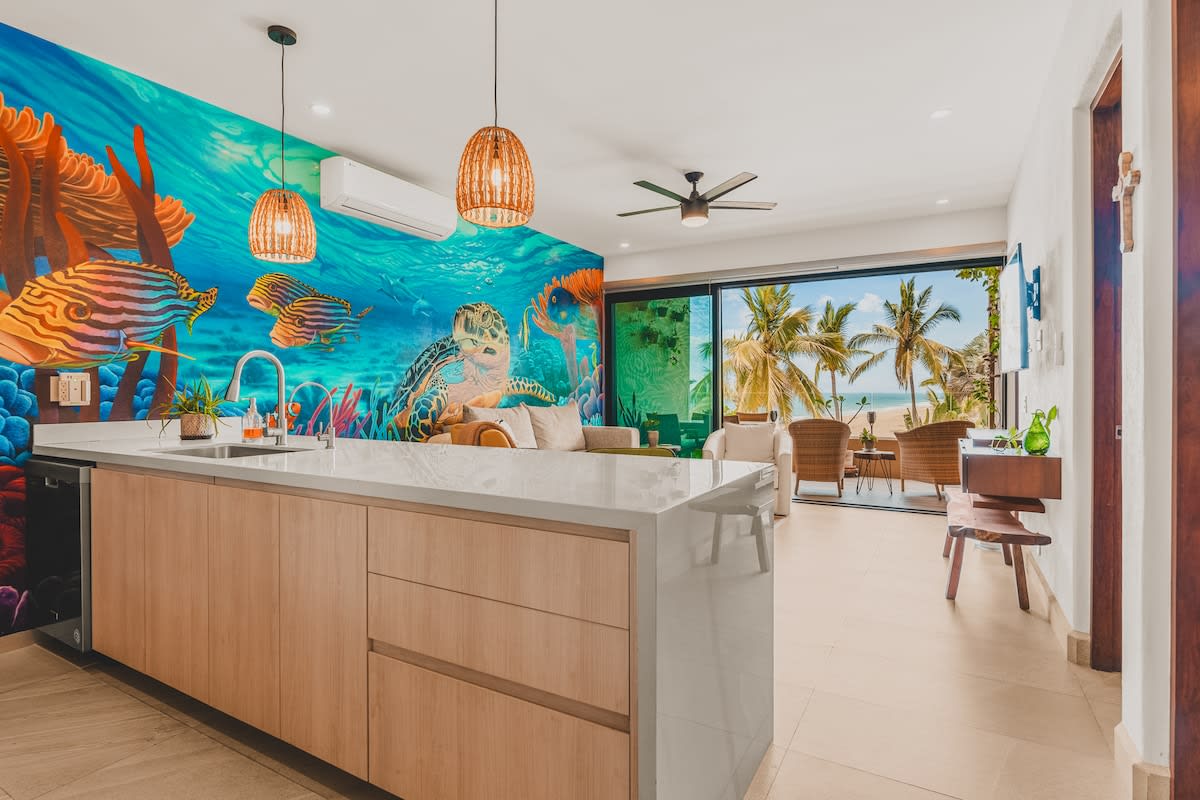 Open-concept kitchen and living area with a large white island, light wood cabinetry, and a striking underwater mural covering one wall, leading to a balcony with ocean views and palm trees.