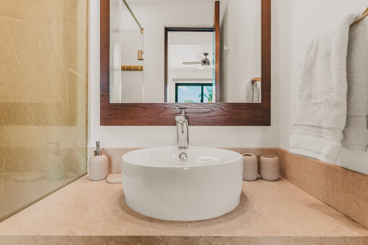 A modern bathroom features a white vessel sink on a light-colored countertop, a wooden-framed mirror, and a chrome faucet. Towels are neatly hung on the left.
