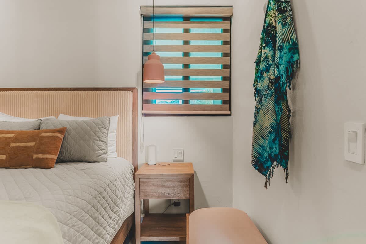 A close-up of the nightstand in bedroom 2 reveals a wooden table with a modern orange pendant light and a white smart speaker. A window with horizontal blinds is visible, and a colorful patterned throw hangs on the adjacent wall as decorative art.